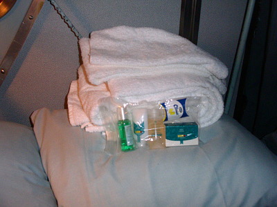 [The shower kit rest against the folded white towels. Kit includes soap, shampoo, rinse, mouthwash, and earplugs. Bedding is a shade of green.]